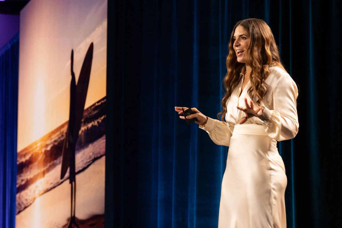 Woman in white dress presenting on stage, gesturing while holding a clicker, blue curtain backdrop.