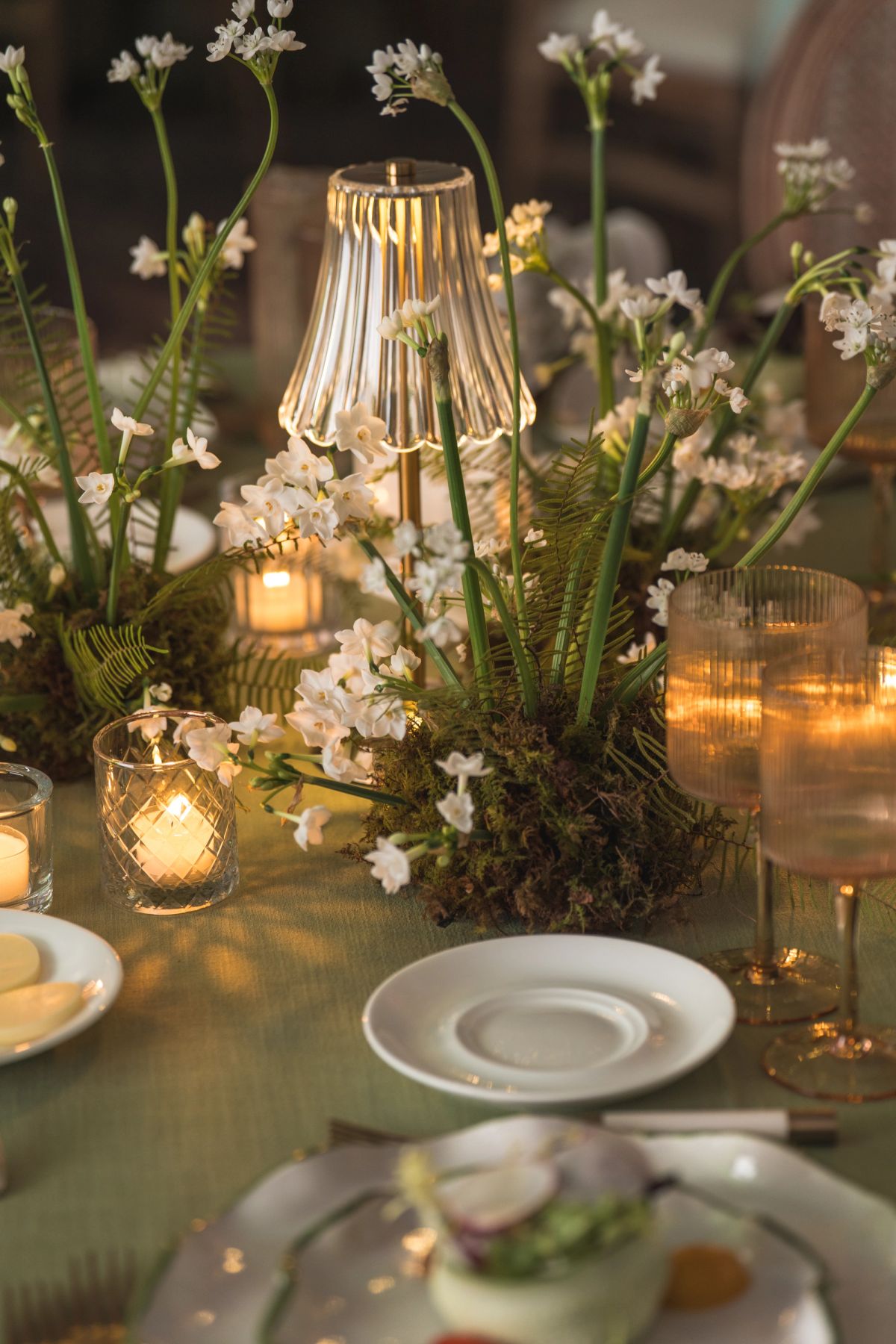 Elegant table setting with white flowers, glowing candles, ribbed glass lamp, and white plates on green.