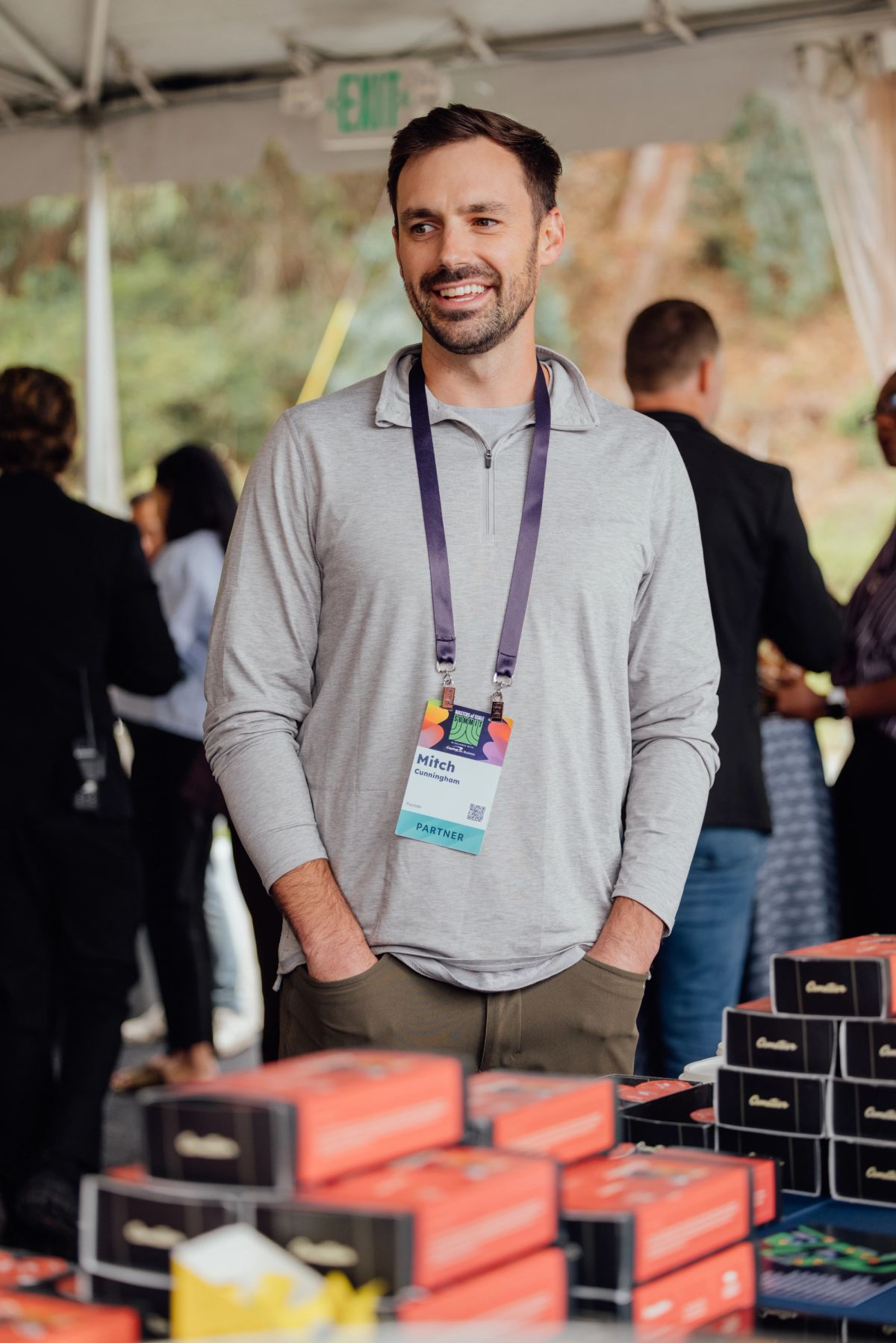 Man wearing conference badge smiles at indoor event with product displays and attendees in background