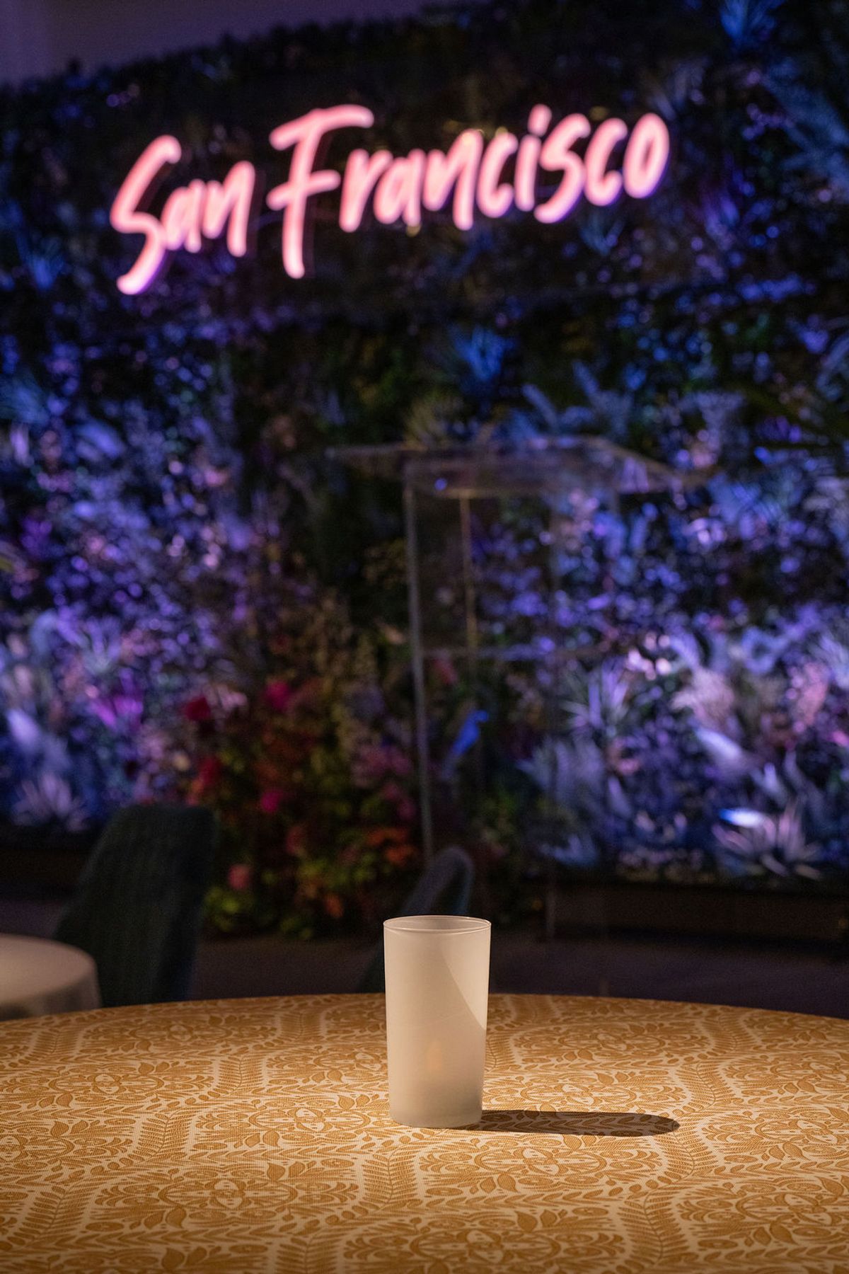 White glass sits on patterned table at evening reception with San Francisco neon sign and purple lighting.