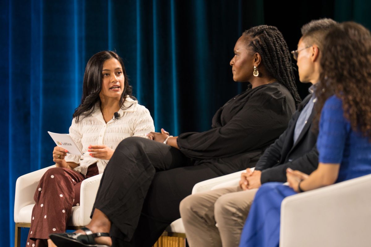 Three women in discussion on a conference panel, seated in white chairs against a blue curtain backdrop.