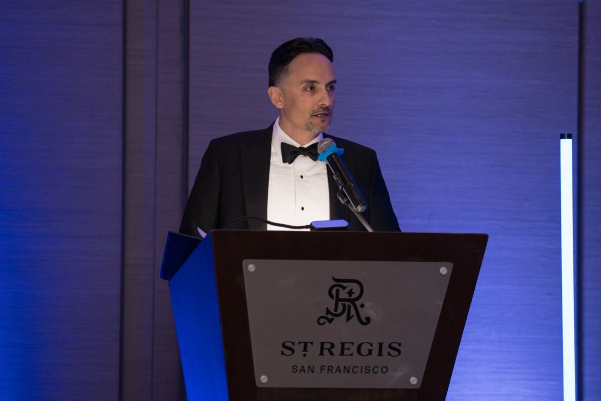 Speaker in tuxedo at podium with microphone at formal gala event with purple lighting and St. Regis branding