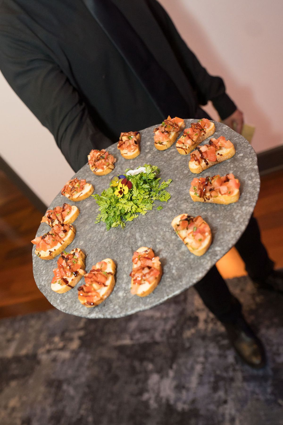 Server in black jacket holding slate platter of bruschetta appetizers with tomatoes and herbs at reception