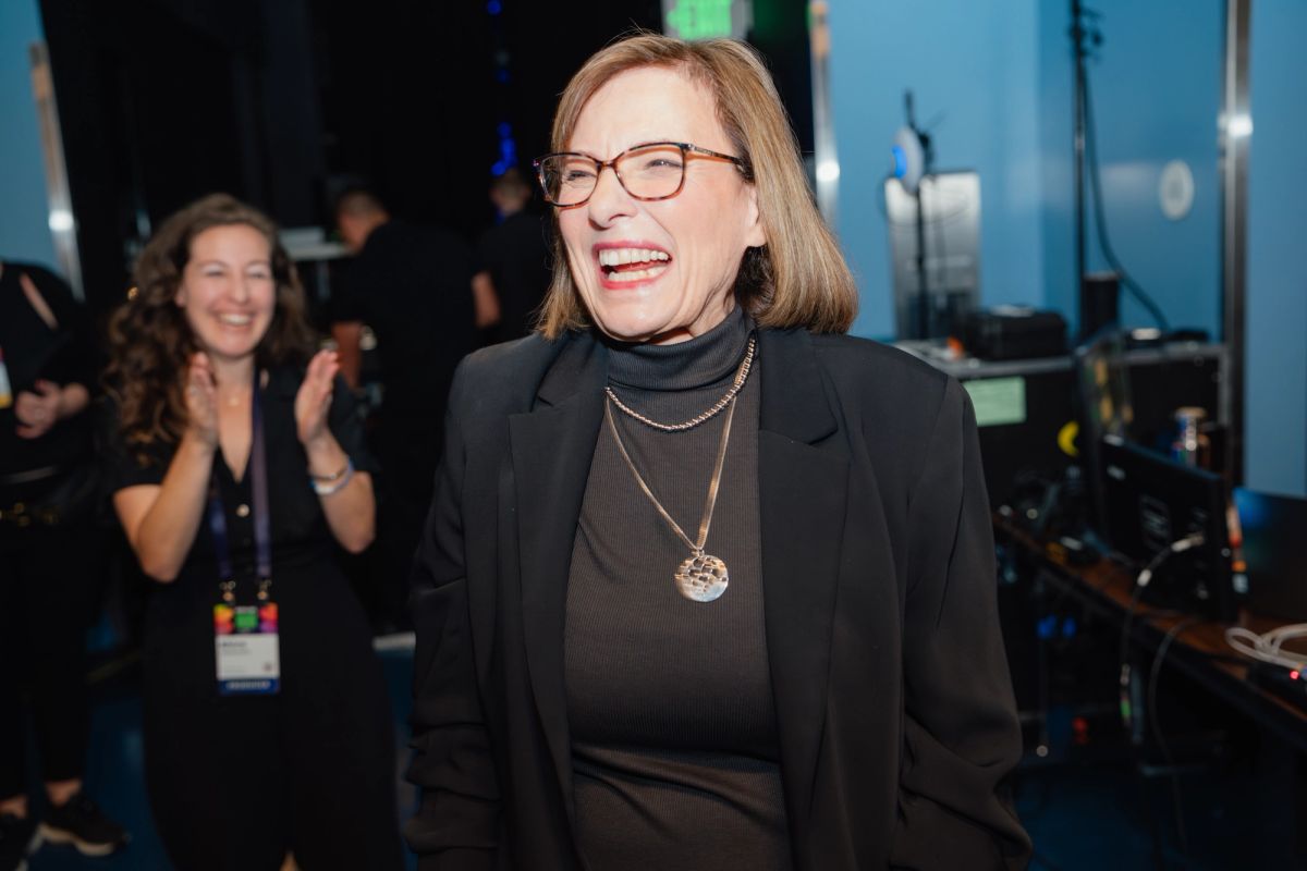 Woman in black blazer and glasses smiling at conference event with applauding attendee in background