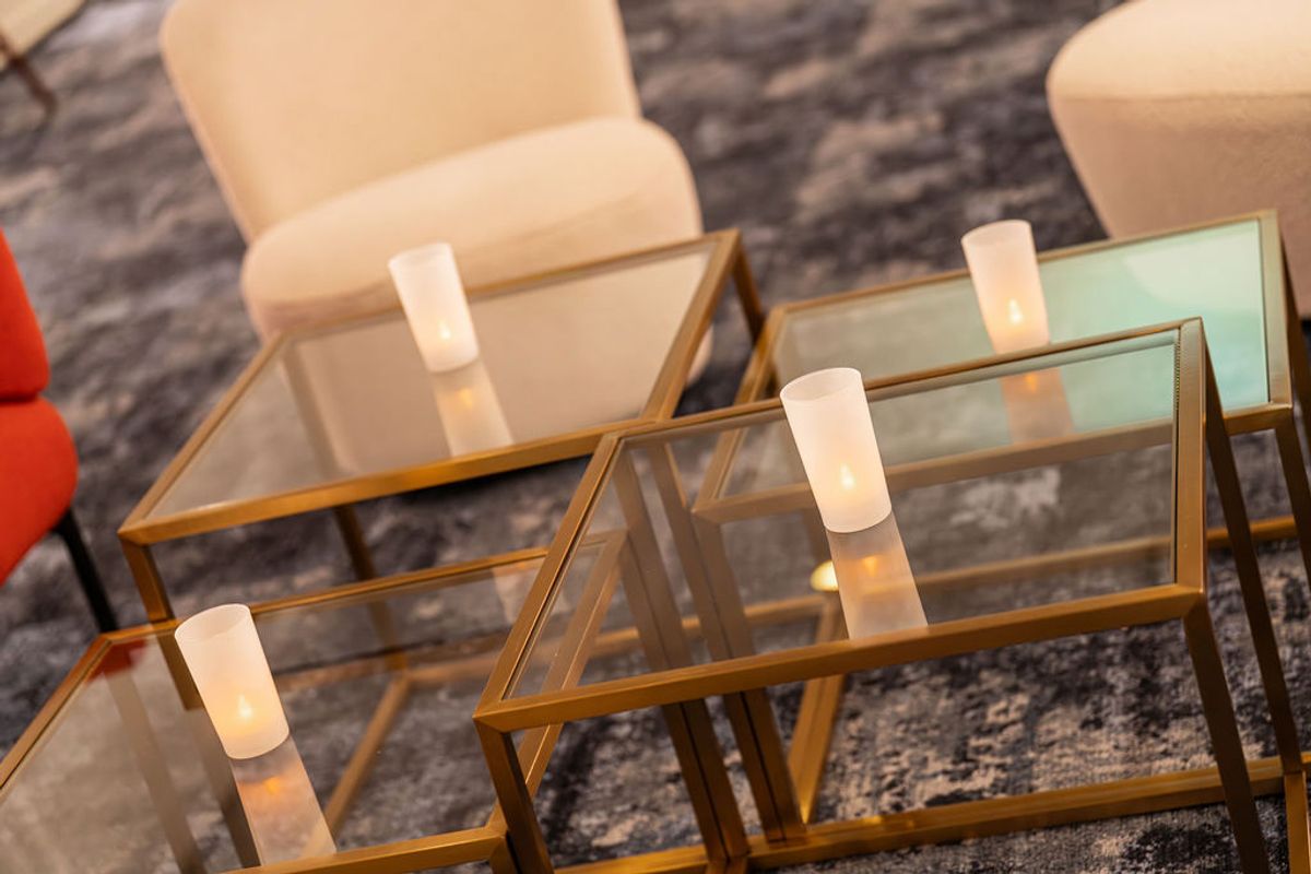 Modern glass and metal side tables with white cylindrical lamps beside beige upholstered chairs on patterned.