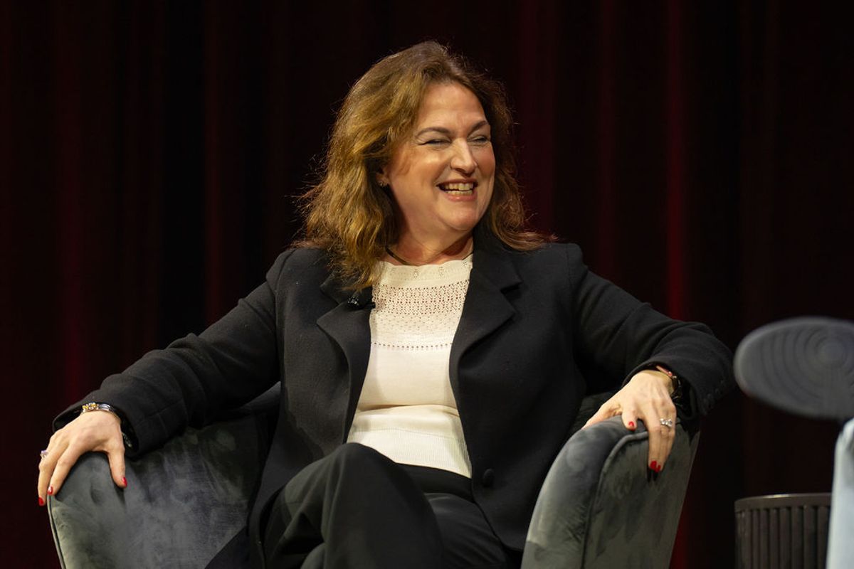 Woman in dark blazer and white top smiling while seated in gray chair during conference presentation