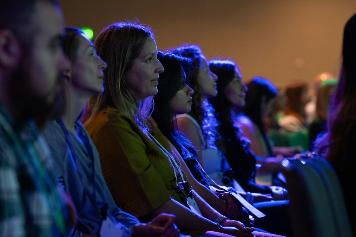 Attendees seated in rows, listening attentively during a conference session in a dimly lit venue.