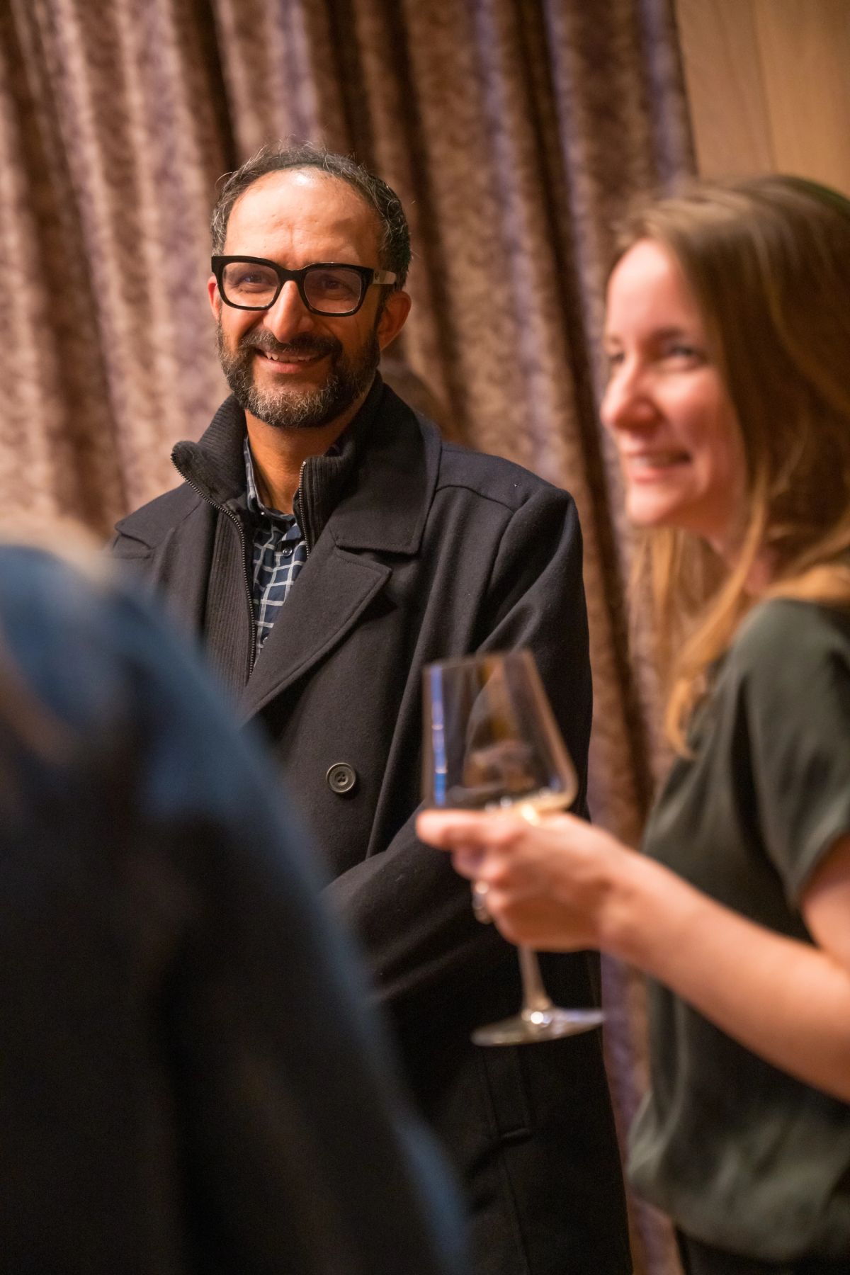 Man in glasses and dark coat smiles while woman holds wine glass at indoor networking event