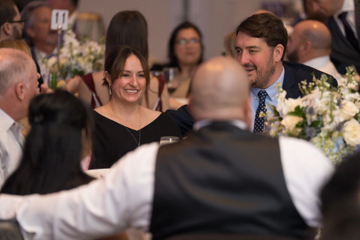 Attendees in formal attire socializing at an elegant dinner event with floral centerpieces and table settings.