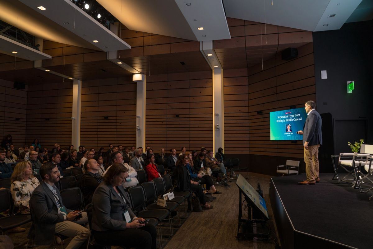 Speaker presenting to seated audience in modern auditorium with wood paneling and projection screen