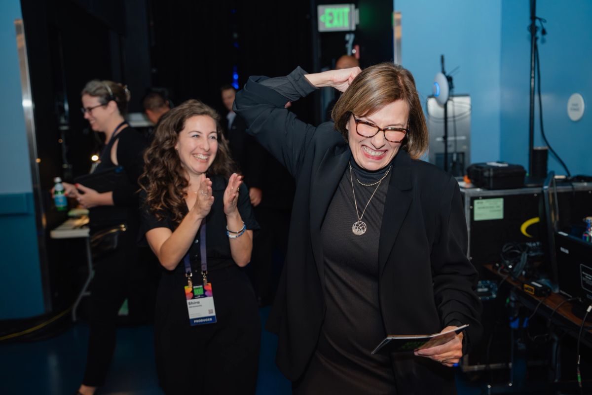 Two women celebrate at a conference, one applauding while the other smiles and holds papers in a blue-walled.