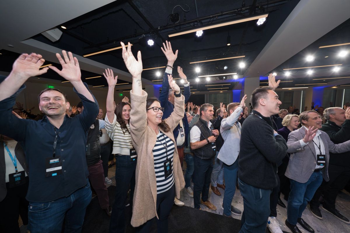 Conference attendees celebrate with raised hands and applause in a modern venue with exposed ceiling.