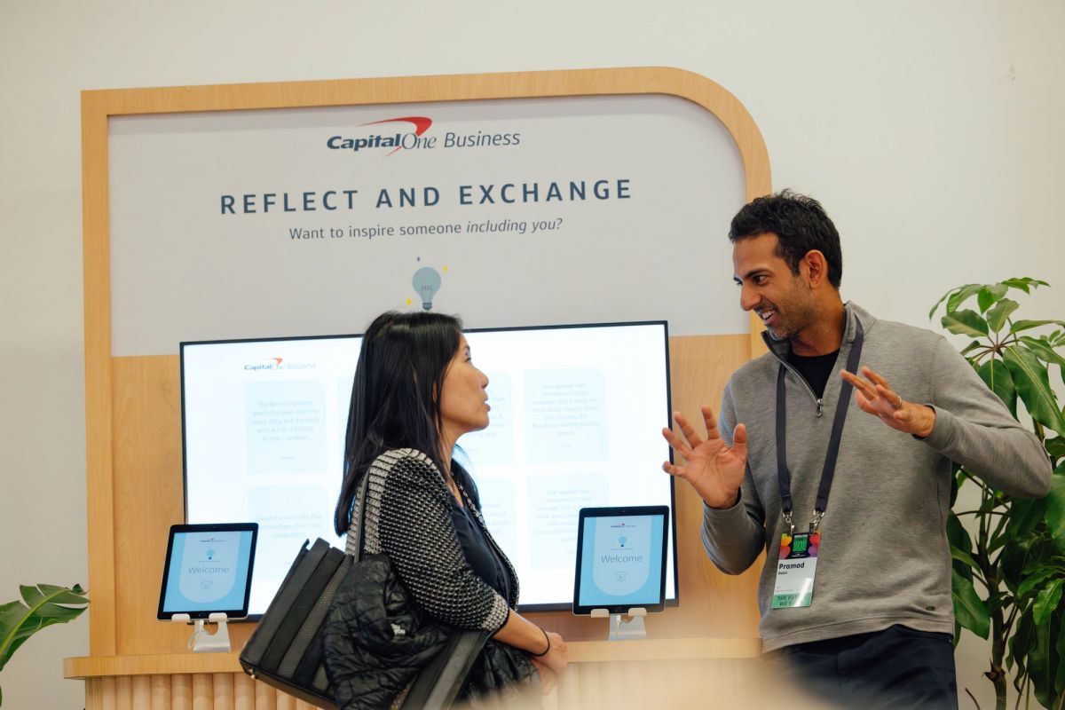 Two people conversing at a Capital One Business booth with "Reflect and Exchange" signage and computer.
