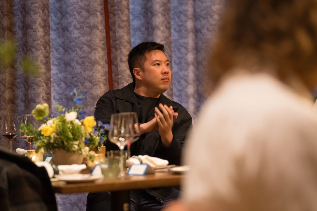 Panelist in black shirt gesturing while speaking at table during discussion, with flowers and wine glasses.