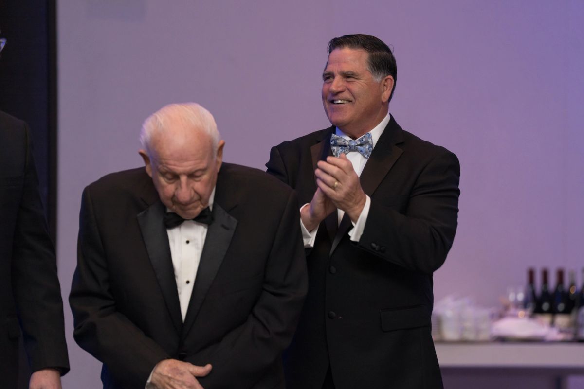Two men in tuxedos at formal event, one clapping while other looks down, purple backdrop visible