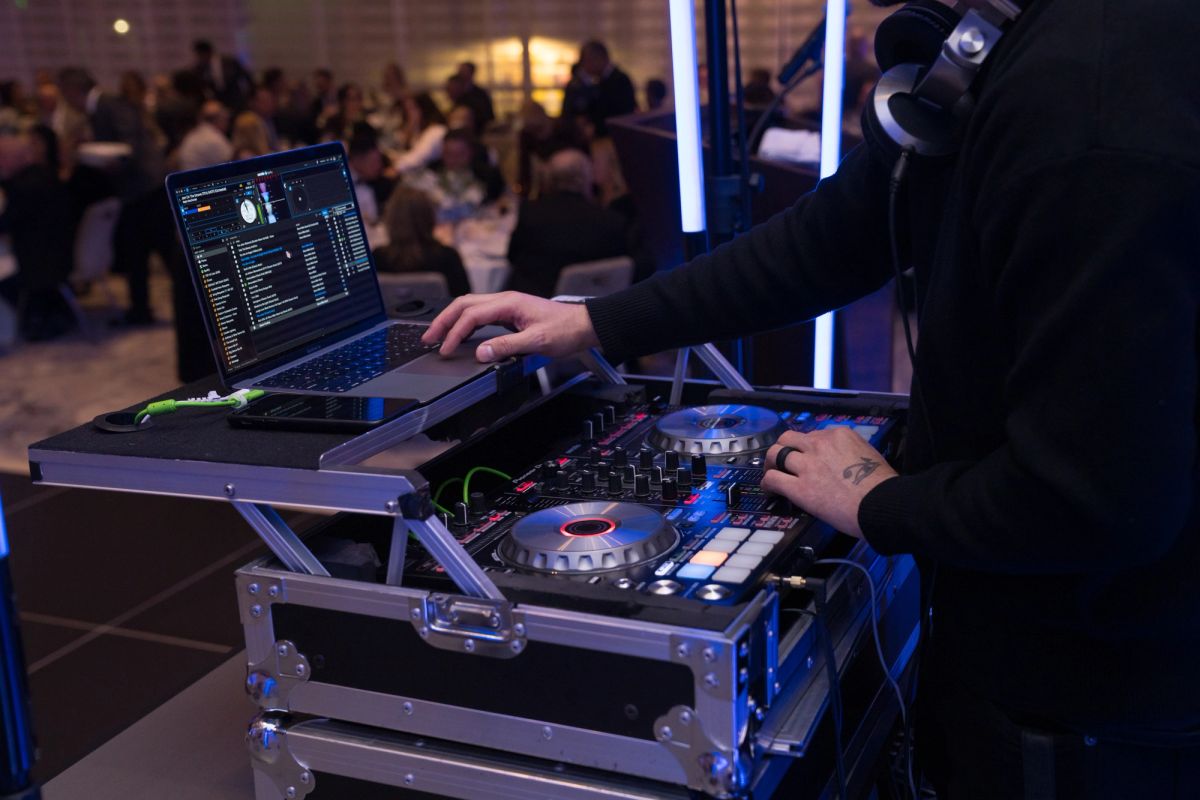 DJ operating mixing equipment and laptop at evening event with crowd of attendees in background