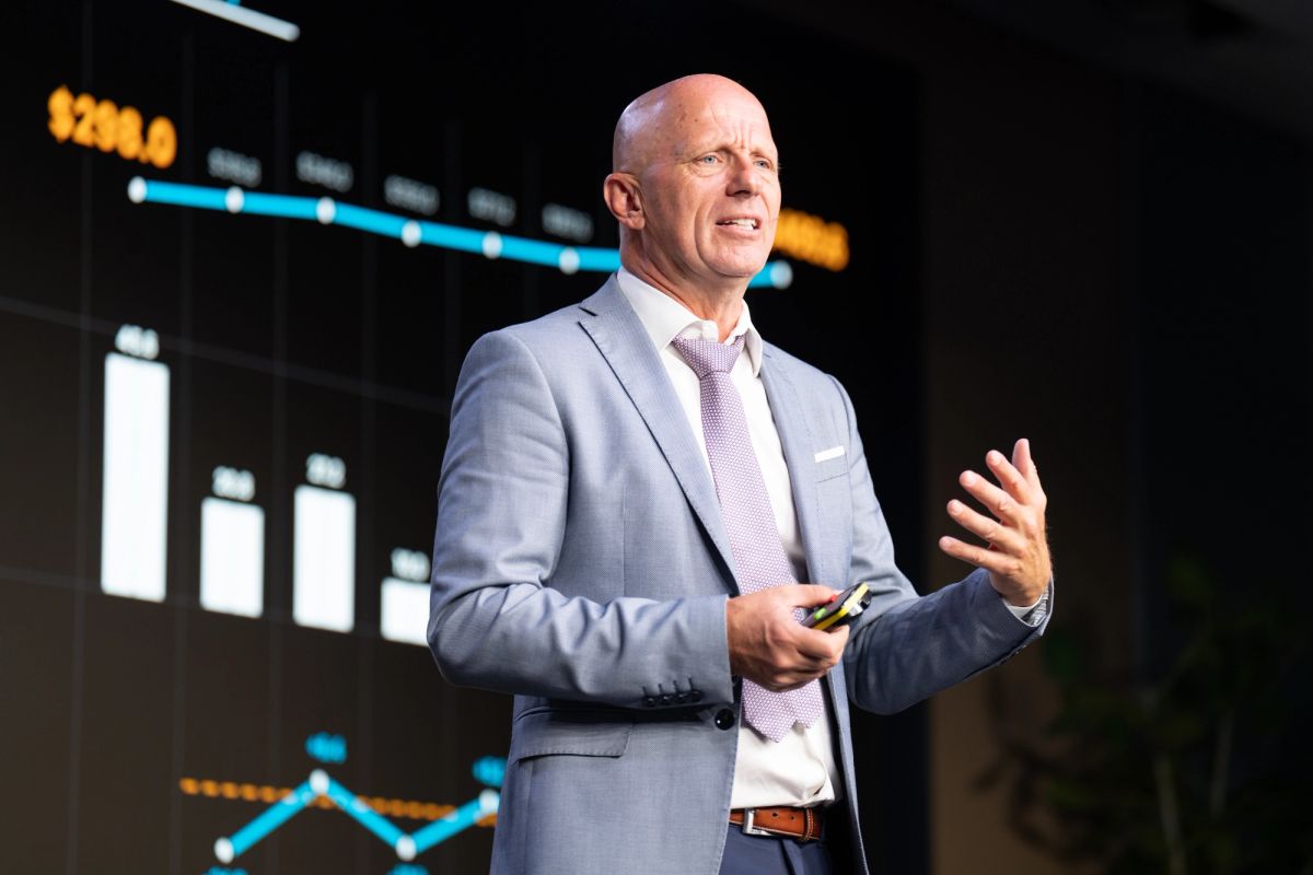 Bald executive in gray suit presenting on stage with data charts displayed on screens behind him