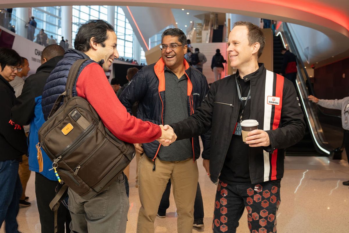 Three men shake hands and chat at a conference networking event in a modern venue with large windows.