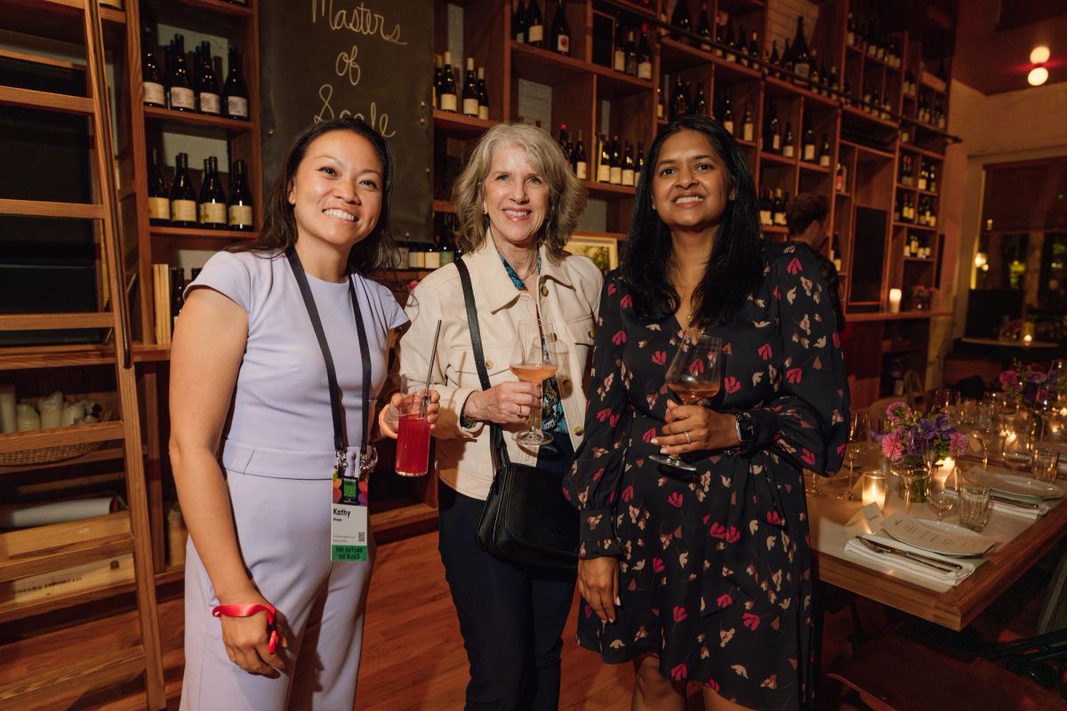 Three women networking with drinks in a wine bar setting with wooden shelves of bottles behind them