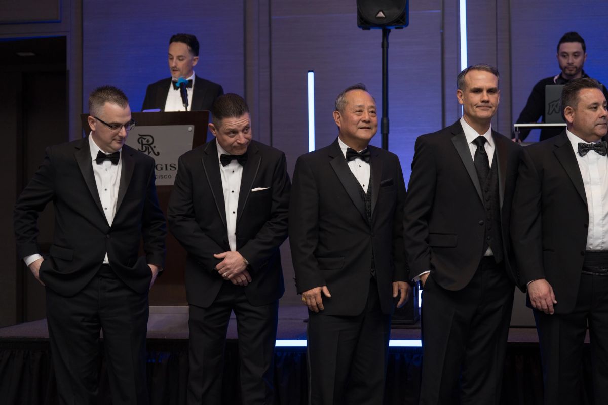 Five men in black tuxedos stand in a line at a formal gala event, with a speaker at podium in background.