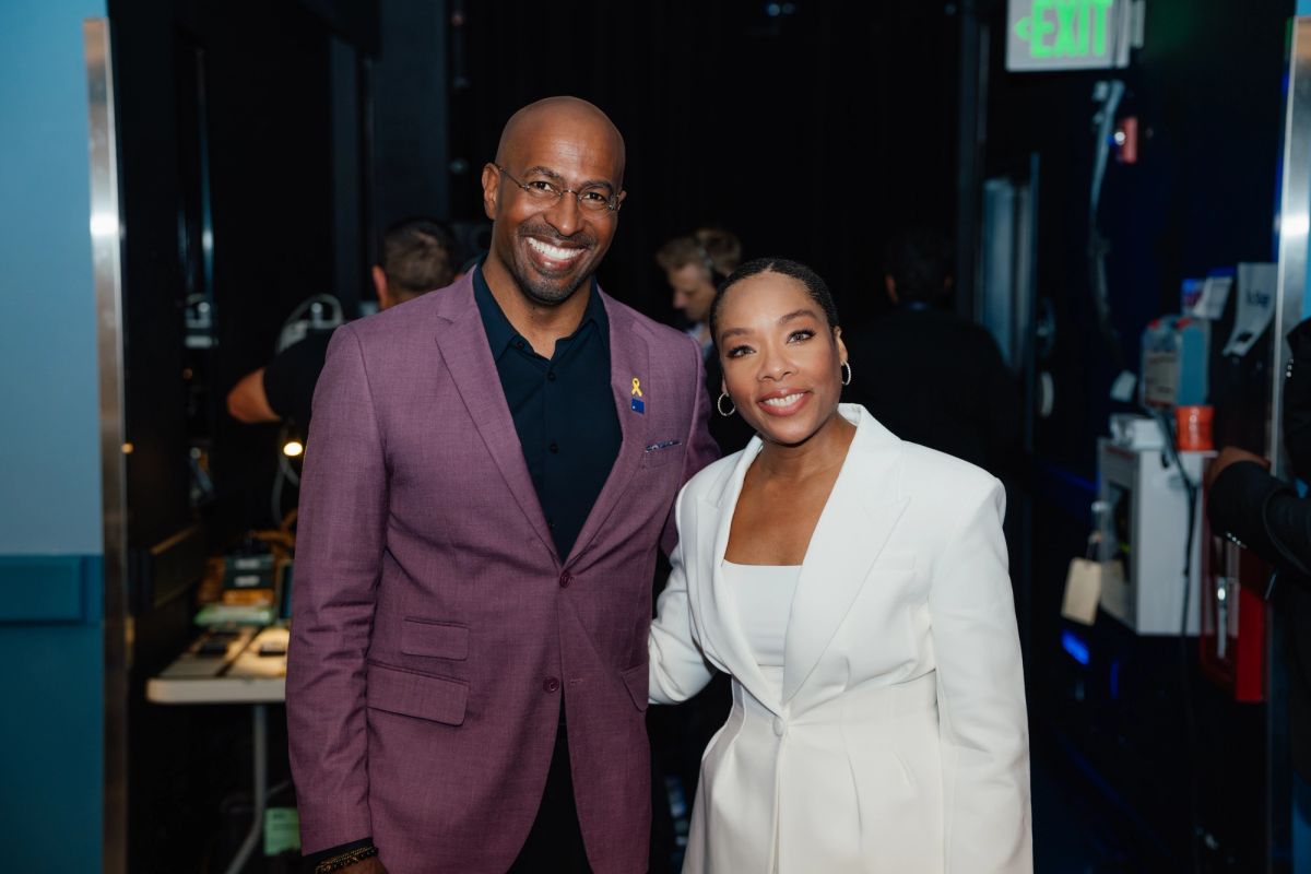 Two people pose together backstage, one wearing a purple suit jacket and one in a white blazer.