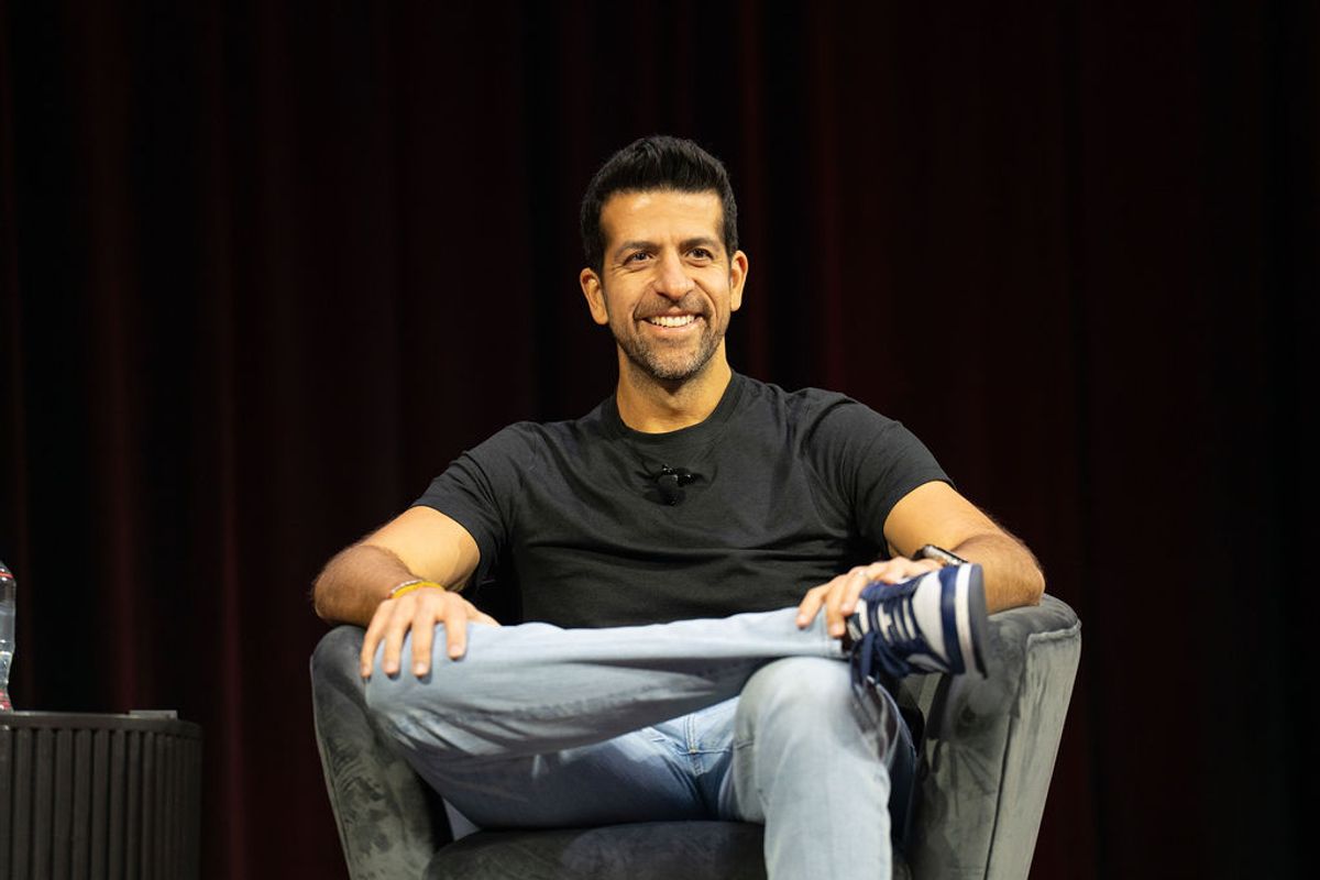 Speaker sitting in gray chair on stage, wearing black shirt and jeans, smiling during interview