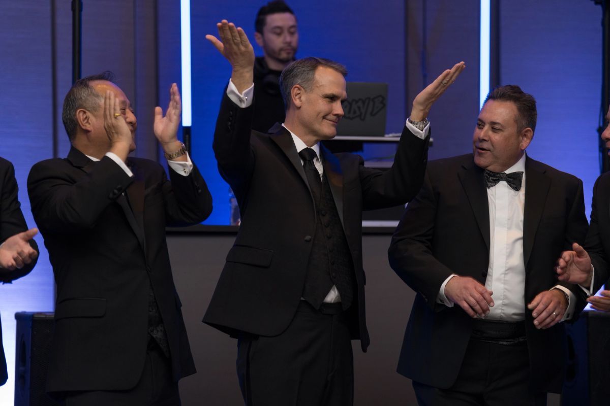 Three men in formal suits celebrating with raised hands at an indoor evening event