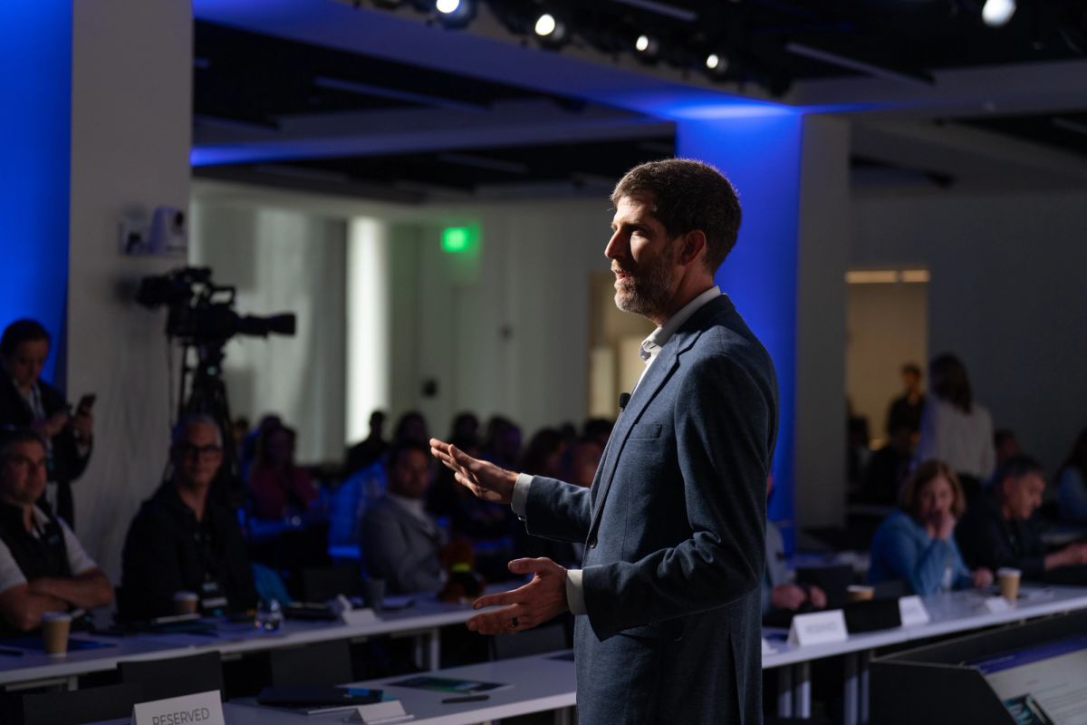 Speaker in gray suit presenting to seated audience in modern conference room with blue lighting