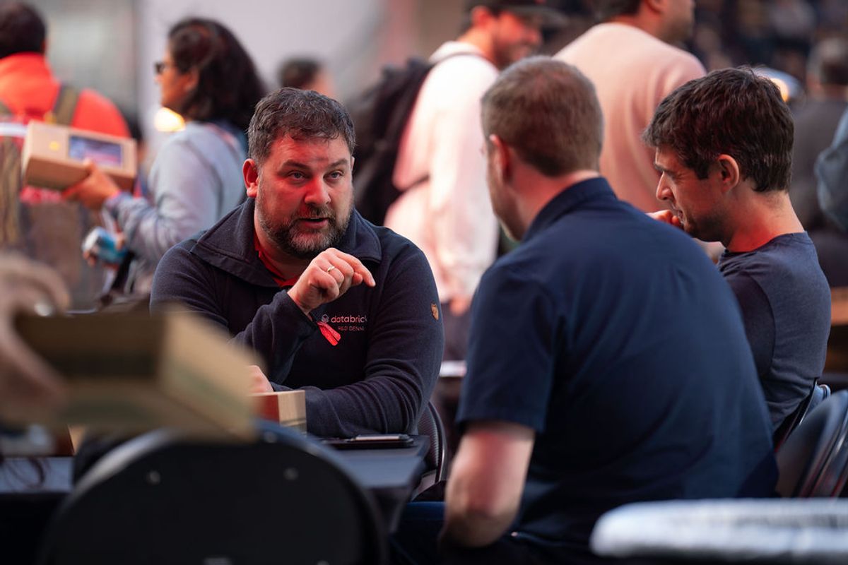 Three men in conversation at a conference networking event with attendees mingling in the background.
