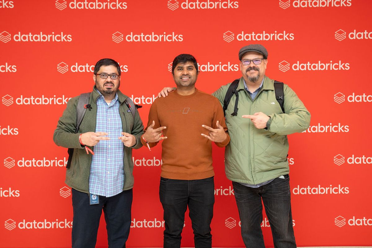 Three men posing together at a Databricks conference, smiling and gesturing at the camera against a red.