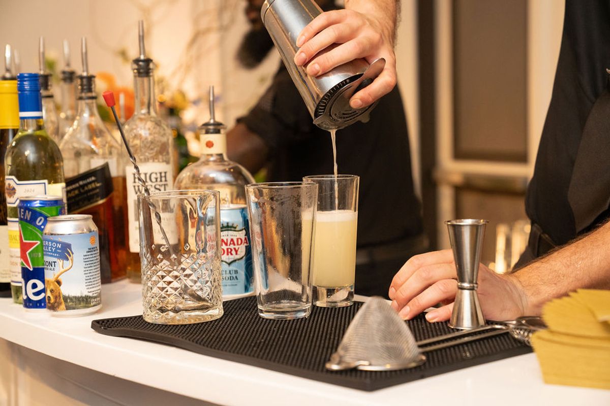 Bartender pours drink from cocktail shaker into glass at bar setup with various liquor bottles and glassware