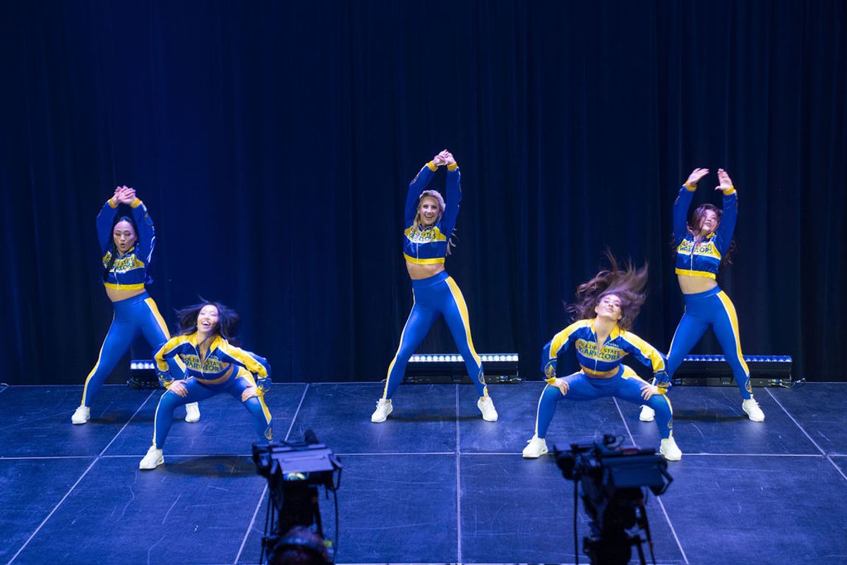 Five cheerleaders in blue and yellow uniforms performing on stage, three standing with pom-poms raised, two.