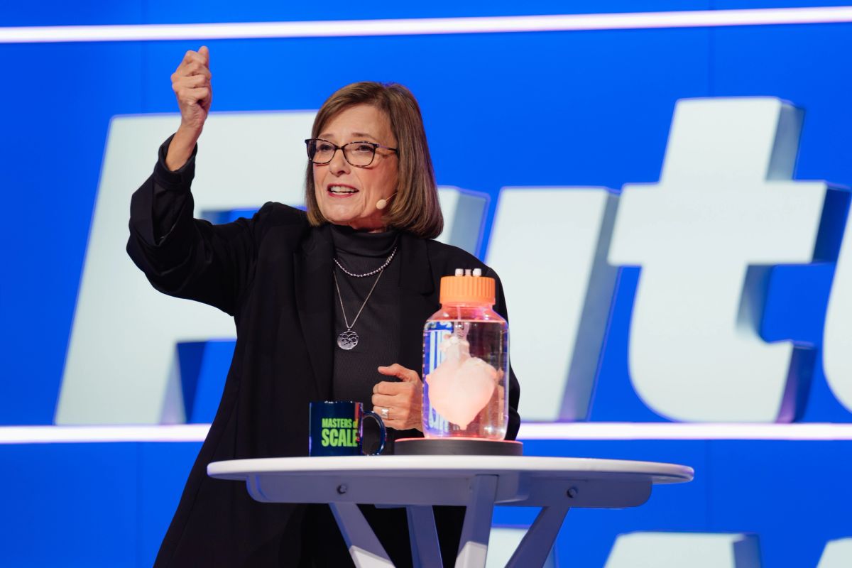 Speaker at podium gesturing with raised fist, holding jar, against blue backdrop with white letters