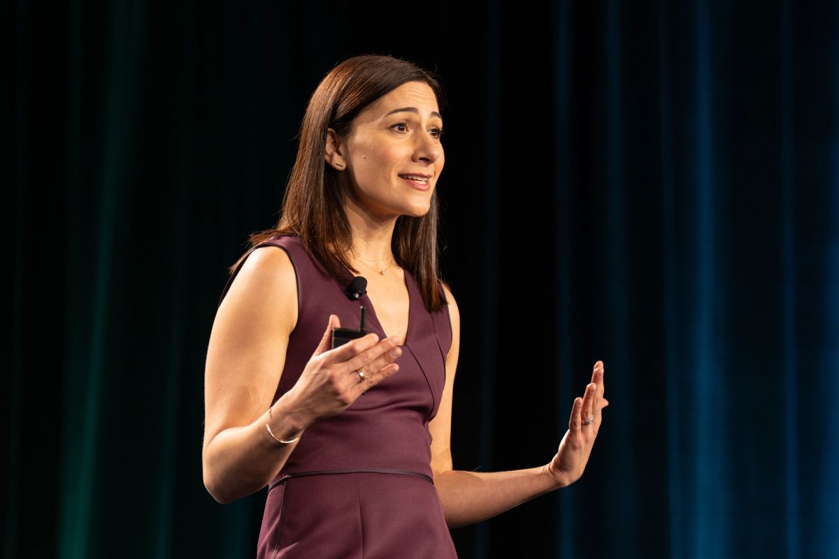 Woman in burgundy dress gestures while speaking on stage at a conference.