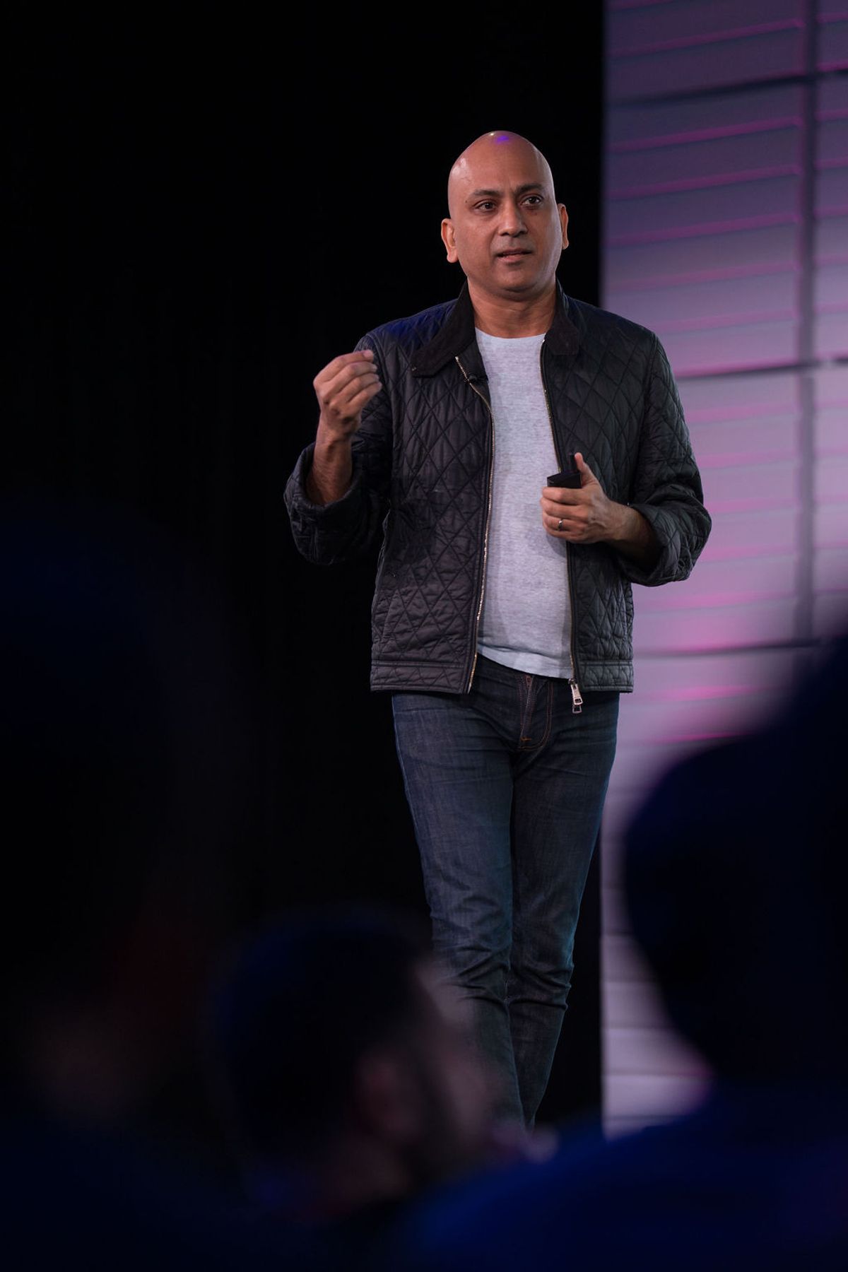 Speaker in black quilted jacket and jeans gesturing while presenting on stage with purple lighting