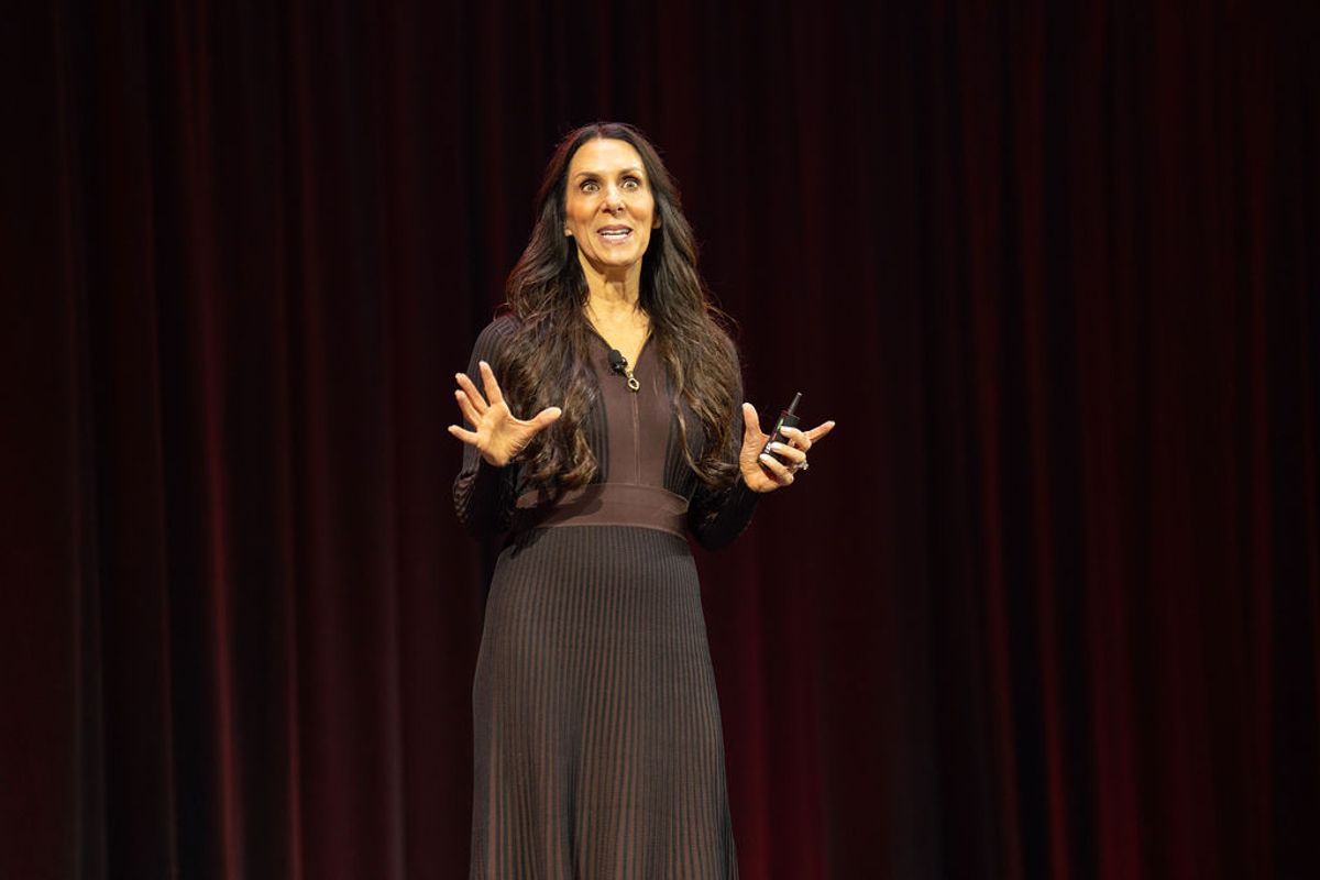 Speaker presenting on stage with hands gesturing, wearing dark outfit against red curtain backdrop