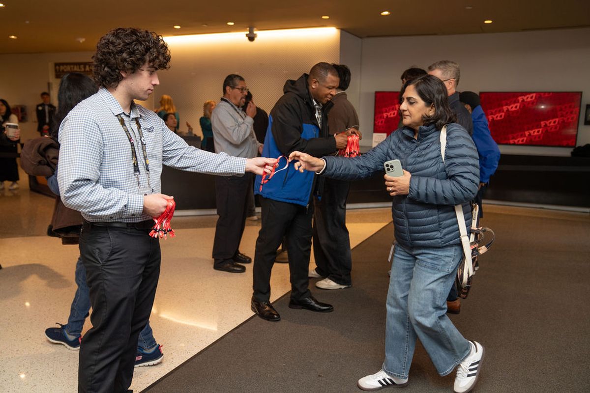 Conference attendees exchange gift bags in a modern lobby with red display screens in the background.