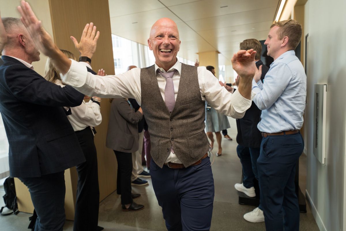 Smiling man in vest walks through office hallway while colleagues give high-fives in celebration