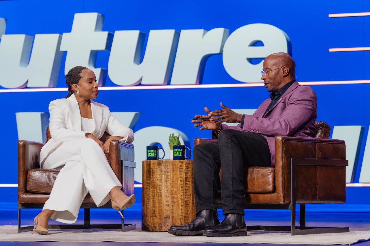 Two speakers in conversation on stage, seated in leather chairs with a small table between them at a.
