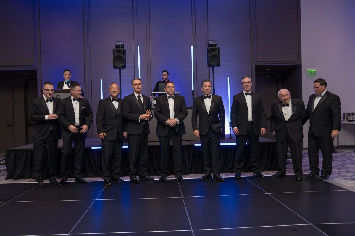 Group of men in formal tuxedos standing on stage at gala event with purple lighting and speakers