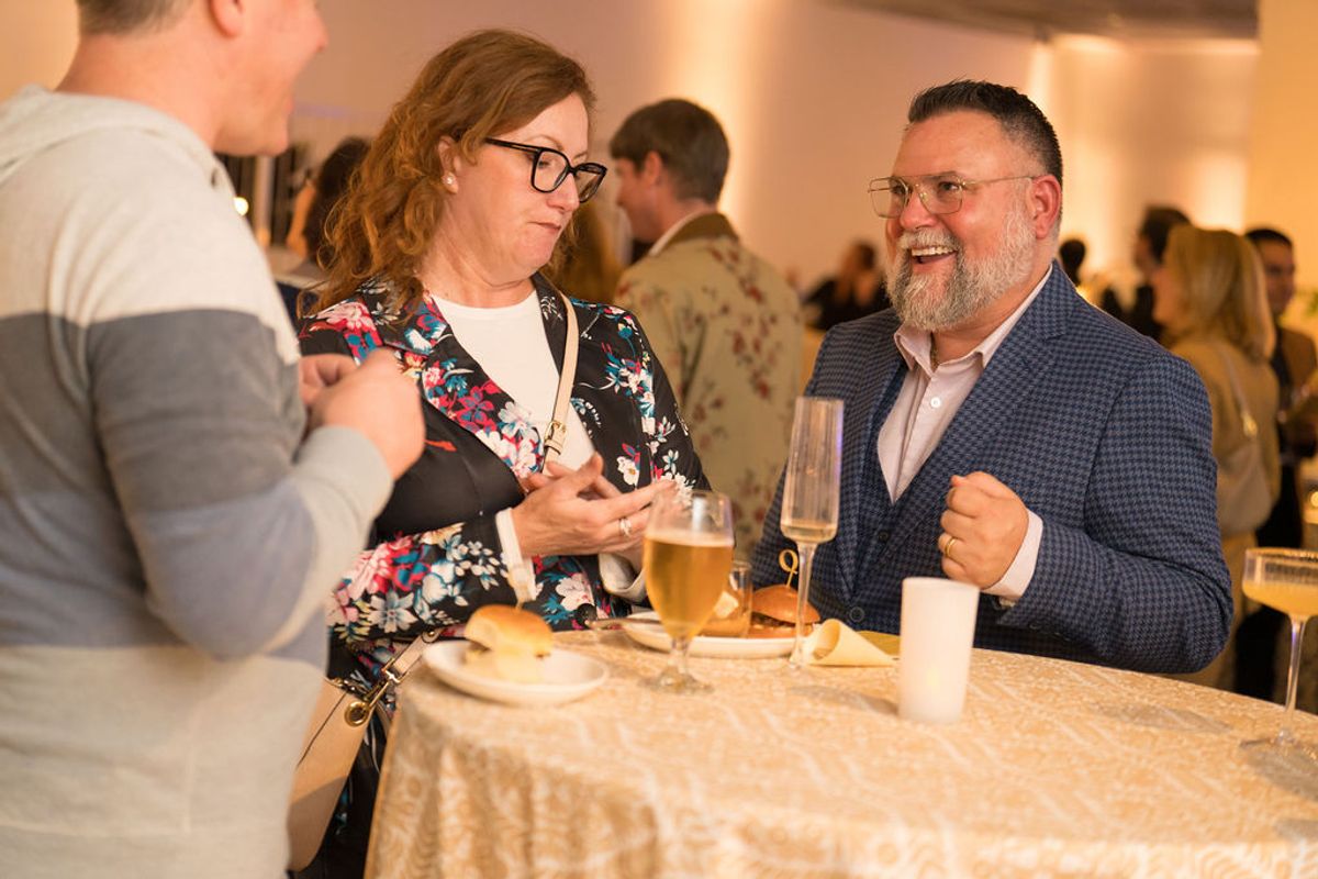 Three professionals network at a cocktail reception, holding drinks and conversing at a high-top table.