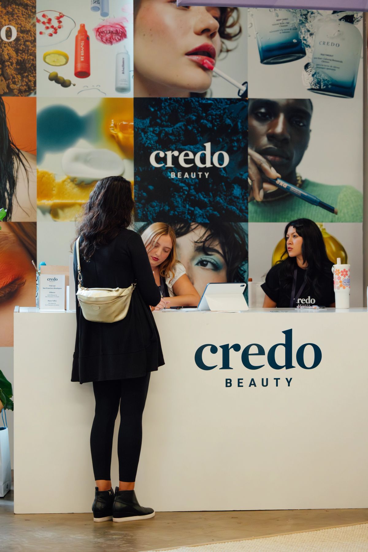 Two people at a Credo Beauty booth, one seated at laptop, another standing nearby with handbag.