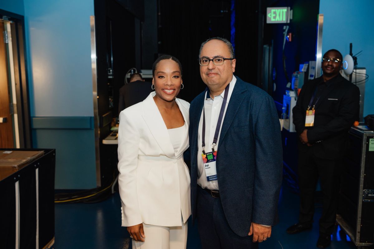 Two professionals pose together backstage at a conference venue, with crew member and exit sign visible in.