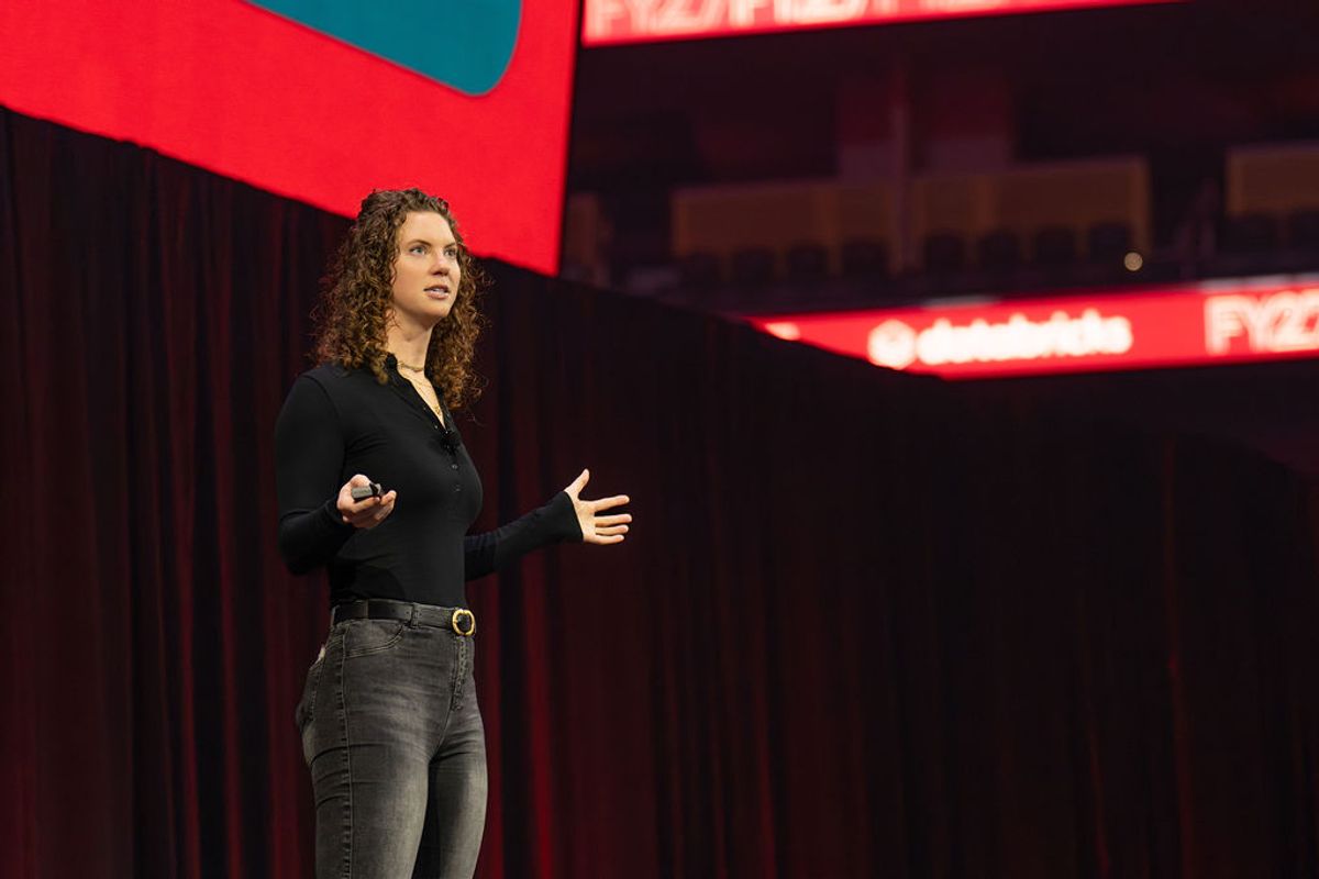 Speaker with curly hair presenting on stage, holding device and gesturing, with red lighting and backdrop