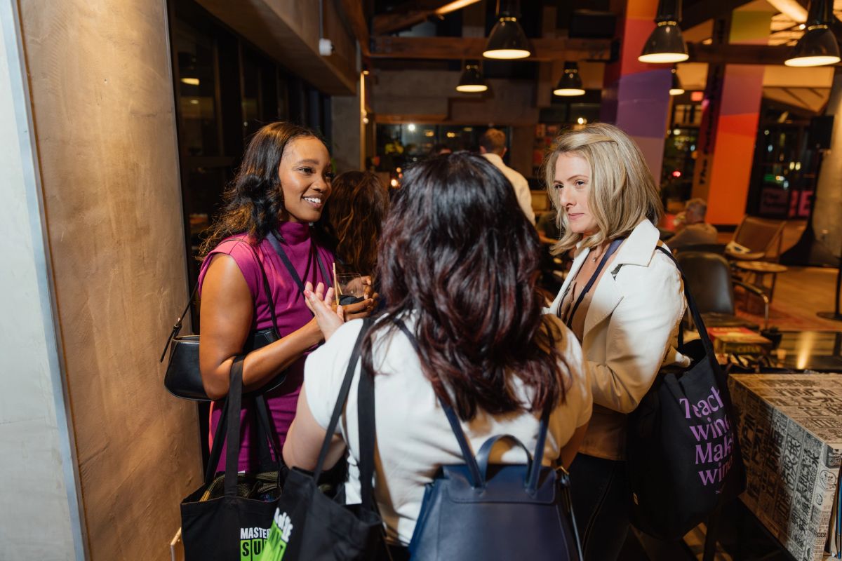 Three women networking and conversing at an indoor conference event with industrial lighting and seating area.