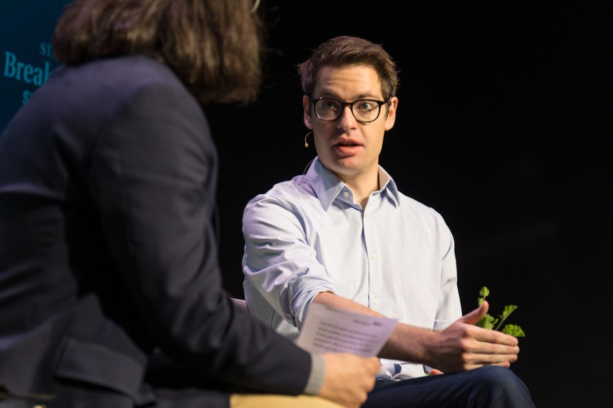 Speaker in glasses and light blue shirt gesturing while speaking to seated interviewer on stage.