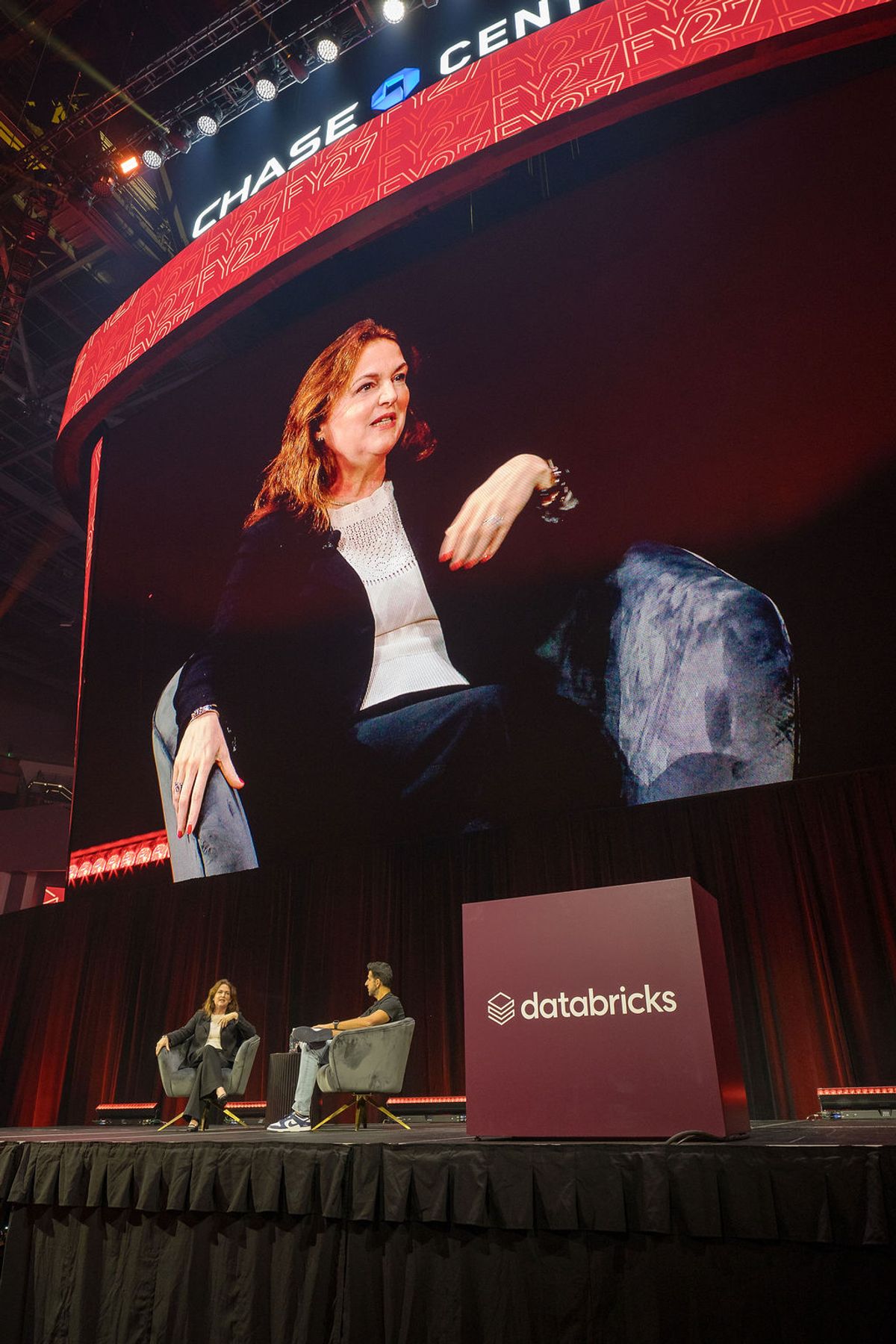 Speaker gesturing during interview on stage at Chase Center with Databricks branding visible on screen