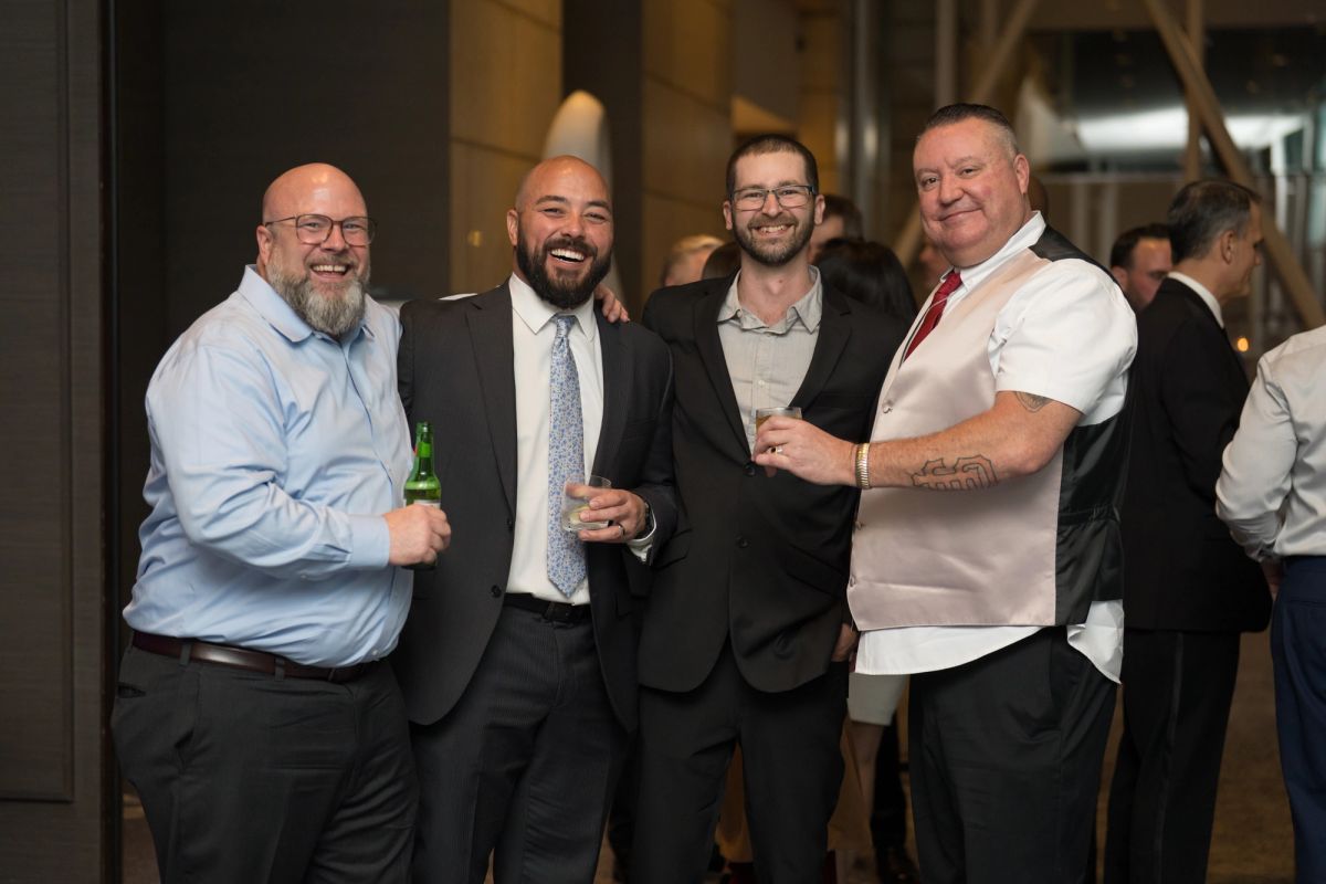 Four men in business attire smiling and holding drinks at an indoor networking event