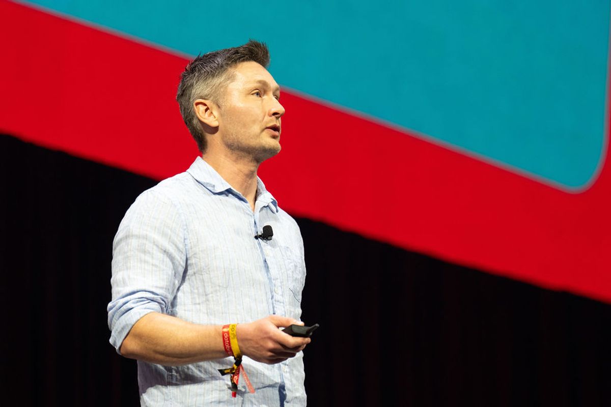 Speaker in light blue shirt raises arm on stage with red and teal backdrop at conference venue