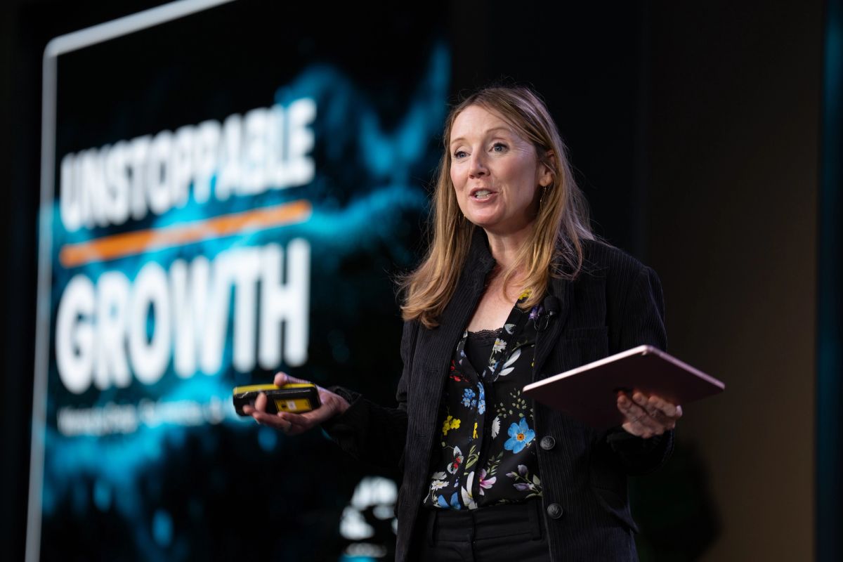 Speaker presenting at conference holding notes and small object with "GROWTH" visible on screen behind her